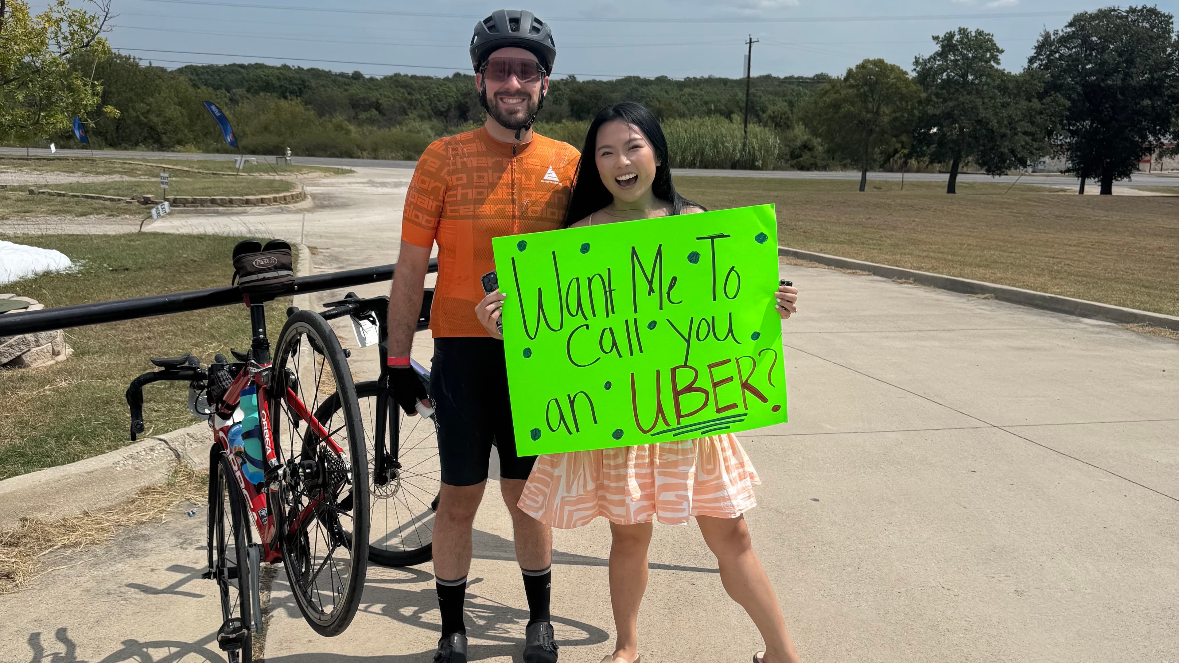 A photo of me in my cycling kit that's orange and black, standing with my wife who holds a sign that reads "Want me to call you an uber?"