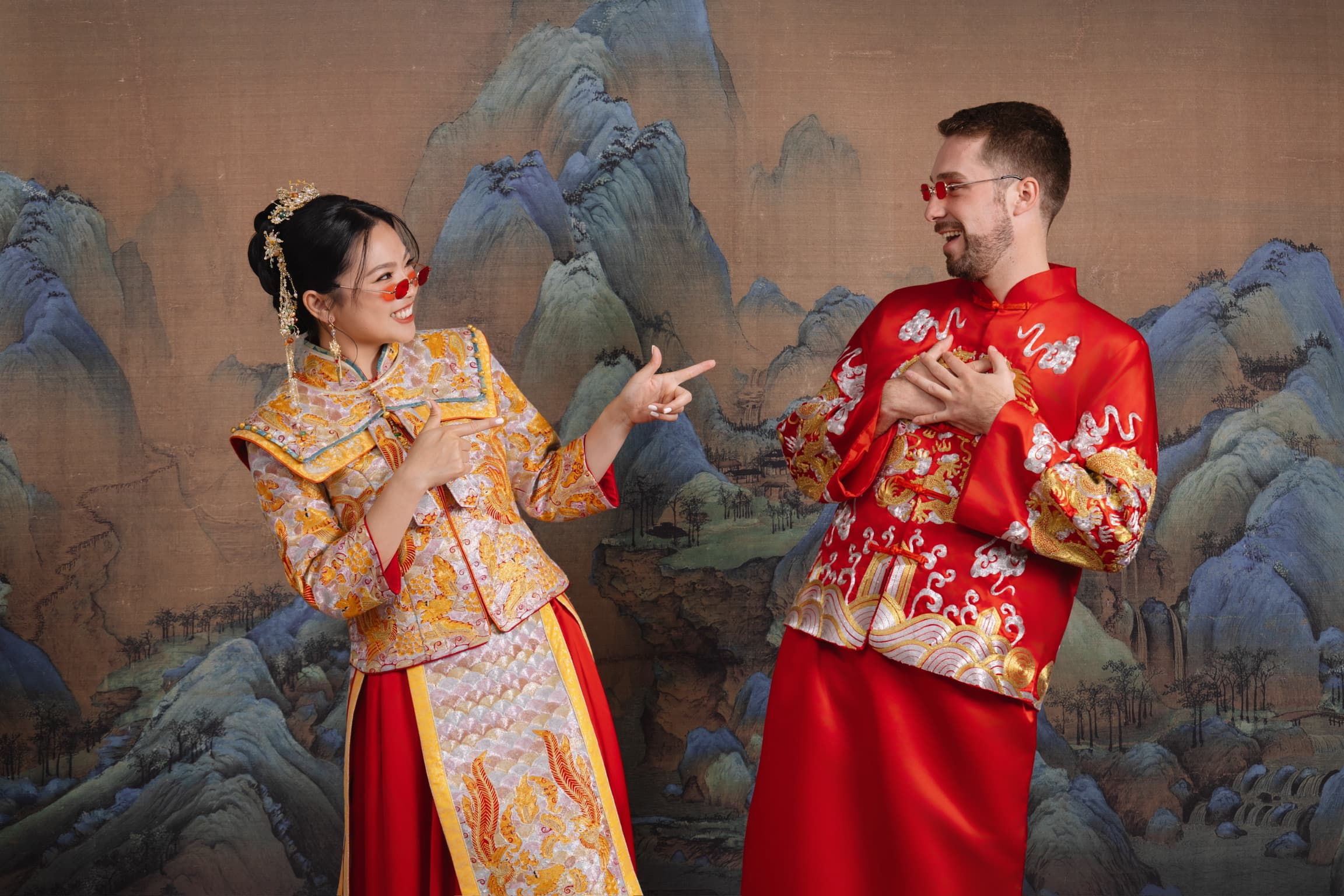 A photo of me and my wife in traditional Chinese red and gold outfits wearing red sunglasses, being a bit goofy and pointing at each other smiling